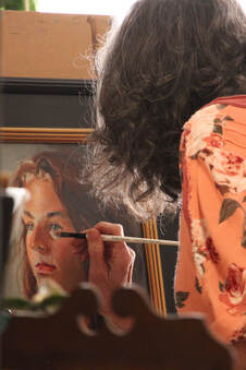 Charlotte Arnold is bent over a framed oil portrait. In her right hand is a fine-tipped paintbrush, poised to add a small stroke under the subject's left eye.