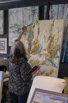 Carol Ann Cain stands at her easel. Her left hand, holding a paintbrush, is raised to paint in the top left corner of a large canvas. The incomplete painting is an expressive nature scene.